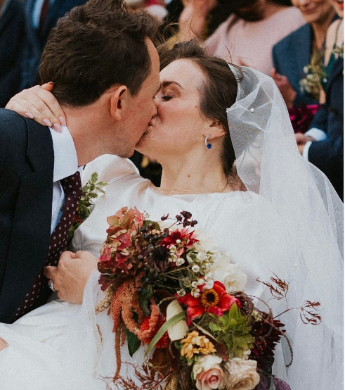 A bride in a white wedding dress and veil kissing her groom, aporting MA tidal hues earrings, with a large autumn-themed floral bouquet in the foreground and blurred guests in the background.
