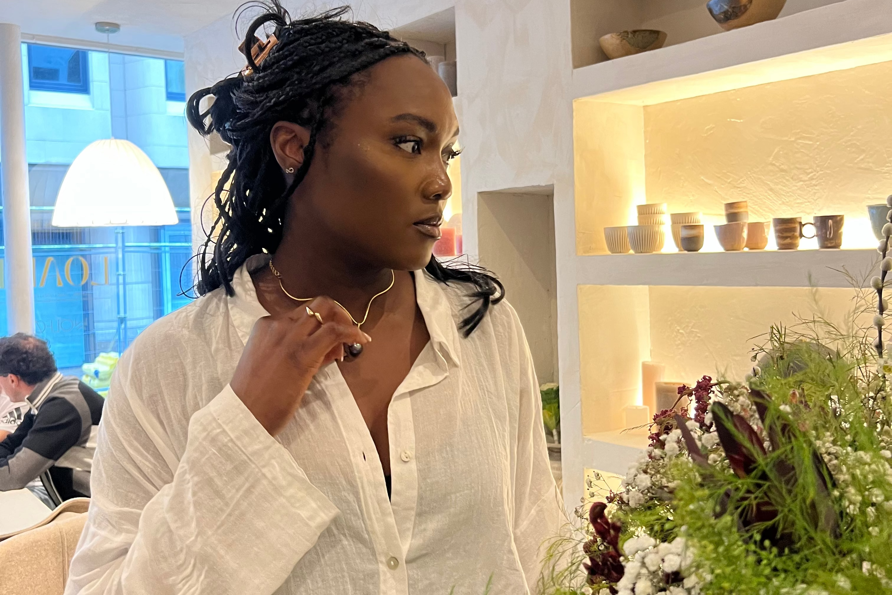 A woman with dark braided hair wearing a white button-up shirt and blue stone pendant stands indoors near a large floral arrangement, with shelves and a softly lit room in the background.