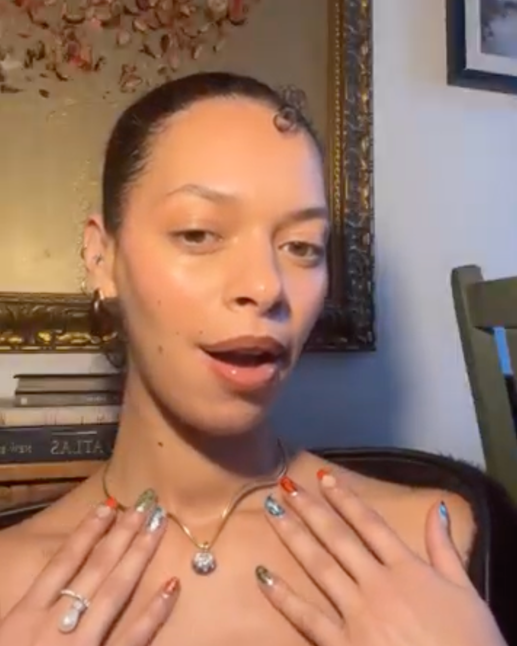 A woman with her hair pulled back is shown from the chest up, wearing a delicate gold necklace with a silver stone and silver hoop earrings. She has silver nail art and a silver ring on one finger, with her hands resting on her chest.